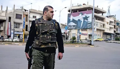 A peine arrivé à Homs, le groupe de journalistes dont fait partie Gilles Jacquier est embarqué vers le rond-point du Gouverneur, à la limite des quartiers Akrama et Al-Nouzha. Un homme en armes veille sur la sécurité des journalistes européens. Derrière lui, une gigantesque affiche du président syrien.
