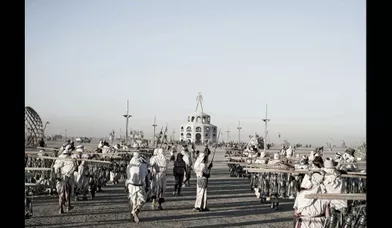 Des bénévoles vêtus de blanc chargés de bouteilles de pétrole vont alimenter les lampadaires. Au fond, la statue de bois monumentale de Burning Man.