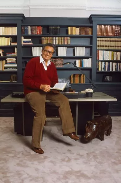 Roger HANIN chez lui dans son appartement du Trocadéro, feuilletant un livre, assis sur son bureau, devant sa bibliothèque.