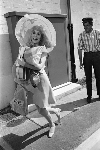 Jacques DEMY coiffé d'une casquette regardant en souriant Catherine DENEUVE en robe et capeline, chantant et dansant des paquets plein les bras avant le tournage d'une scène en extérieur du film "Les demoiselles de Rochefort".