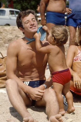 Été 1964, Jean Paul Belmondo en vacances en famille à Beauvallon près de Saint-Tropez. Des vacances bien méritées car l'année 1964 a été pour Jean-Paul un festival de grands rôles qui ont fait de lui une grande star adorée des Français, avec entre autre «L’Homme de Rio», «Week-end à Zuydcoote», et «Cent-mille dollars au soleil». On le voit ici entrain de jouer sur la plage avec son fils Paul.