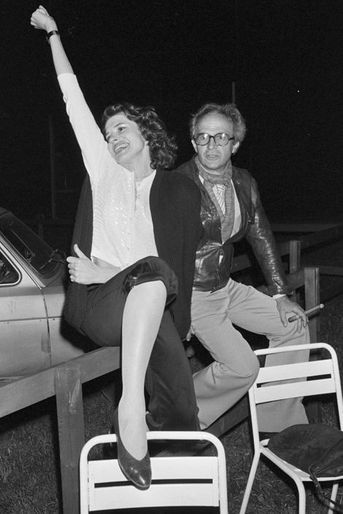 France, Grenoble, 8 mai 1981, pendant le tournage du film "La femme d'à côté", lors d'une soirée de repos, le réalisateur François TRUFFAUT est assis sur une barrière de bois près de l'actrice Fanny ARDANT, à l'époque sa compagne. Elle sourit en tendant un bras en l'air, une jambe sur une chaise en métal posée devant elle. Lui tient un cigare dans une main. Derrière eux, une voiture garée.