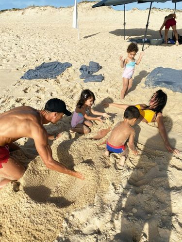 Escale sur la plage de Comporta (Portugal). Georgina est recouverte de sable par les jumeaux Eva et Mateo avec l’aide de Ronaldo, sous les yeux rieurs d’Alana Martina.