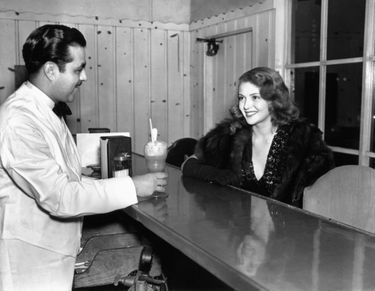 Lana Turner rejoue pour les photographes le moment où elle a a été découverte, en commandant un soda au même serveur, Manuel Hernandez, au ‘Tops' Fountain, en face de son lycée, sur Sunset Boulevard à Hollywood, California, en novembre 1937.