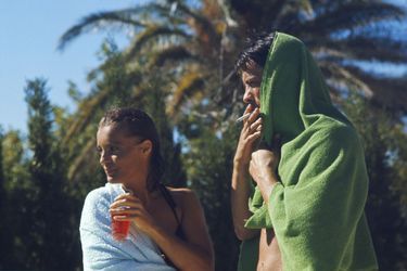 Tournage du film "La piscine" de Jacques DERAY dans le décor d'une somptueuse villa aux environs de Saint-Tropez pourvue d'une immense piscine : plan de trois-quarts de Romy SCHNEIDER un verre à la main aux côtés d'Alain DELON fumant une cigarette, tous deux enroulés dans une serviette lors d'une pause.