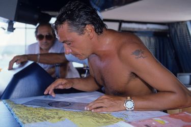 Monte-Carlo, août 1984 : le chanteur Michel Sardou, torse nu, étudie une carte à bord de son bateau.