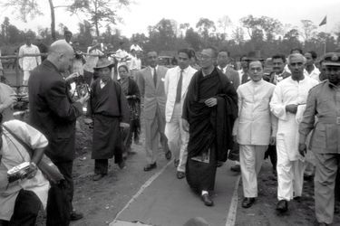 Traqué par les Chinois, le 14e DALAI-LAMA (23 ans) a fui le Tibet et trouvé refuge en Inde : 18 avril 1959 : son arrivée à la frontière de l&#039;Assam, après avoir traversé la région des territoires de la North East Frontier Agency (NEFA) qui sépare l&#039;Inde du Tibet et de la Chine. Avant de rejoindre la ville de Tezpur où lui et ses partisans sont attendus par la presse internationale, le Dalai-Lama fait quelques pas pour saluer le comité d&#039;accueil venu l&#039;attendre à la frontière. A sa gauche, le représentant indien du Pandit Nehru, R. V. MENON.