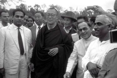 Traqué par les Chinois, le 14e DALAI-LAMA (23 ans) a fui le Tibet et trouvé refuge en Inde : 18 avril 1959 : son arrivée à la frontière de l&#039;Assam, après avoir traversé la région des territoires de la North East Frontier Agency (NEFA) qui sépare l&#039;Inde du Tibet et de la Chine. Avant de rejoindre la ville de Tezpur où lui et ses partisans sont attendus par la presse internationale, le Dalai-Lama fait quelques pas pour saluer le comité d&#039;accueil venu l&#039;attendre à la frontière. A sa gauche, le représentant indien de Nehru, R. V. MENON, et derrière, coiffé de son chapeau, Gyalo THONDUP, un des frères aînés du Dalai-Lama.