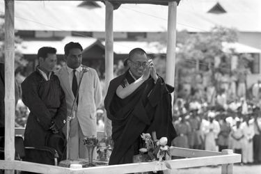 Traqué par les Chinois, le 14e DALAI-LAMA (23 ans) a fui le Tibet et trouvé refuge en Inde. Après avoir traversé la région des territoires de la North East Frontier Agency (NEFA) qui sépare l&#039;Inde du Tibet et de la Chine, le Dalai-Lama à fait son entrée dans la ville de Tezpur en Assam, où lui et ses partisans sont attendus par la presse internationale. De là, il devra prendre le train spécial envoyé par le Pandit Nehru (1er ministre indien) pour atteindre sa résidence définitive, à Mussoorie. Tezpur, 18 avril 1959 : sous un toit de chaume tibétain, le Dalai-Lama salue la foule des fidèles qui se sont rassemblés pour écouter et recevoir la bénédiction du &quot;Bouddha vivant&quot;. En arrière-plan à gauche, Gyalo THONDUP, un des frères aînés du Dalai-Lama.