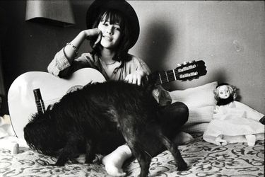 Attitude souriante de Sophie Marceau coiffée d&#039;un chapeau, assise en tailleur sur son lit, entourée de sa guitare, de son chien Rubens un schnauzer nain et d&#039;une poupée chez ses parents à Gentilly.
