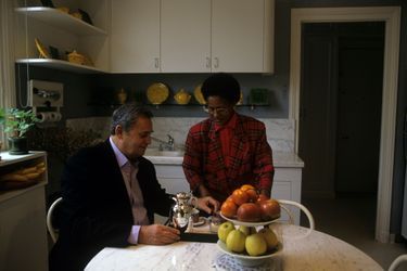 1996. Roger HANIN chez lui, assis dans la cuisine devant une femme préparant le plateau pour le café