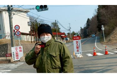 <br />
Vendredi 1er avril, à Miyakoji Hiroki, 28 ans, montre les pastilles d’iode qu’il doit avaler en cas d’accident grave. Sur son visage, un simple masque antipollution. A l’arrière-plan, deux policiers en tenue de protection blanche inspectent un véhicule.