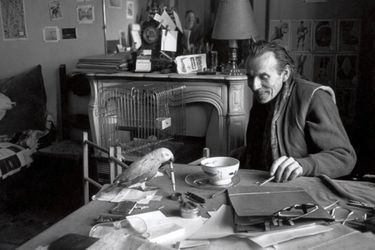 Louis-Ferdinand Céline en 1957 dans sa maison de Meudon. L’écrivain, alors âgé de 63 ans, est assis devant un bol de café posé sur la table sur laquelle se trouve son perroquet Toto avec un stylo dans le bec. 